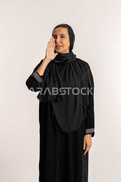 سيدة سعودية تقوم بالنداء بصوت منخفض والكلام بصوت عالي، مفهوم الدردشات والمحادثات، بورتريه مقرب لامرأة عربية خليجية إماراتية محجبة ترتدي عباءة وردية تضع يدها على فمها بإيماءات تدل على المناداة، خلفية بيضاء