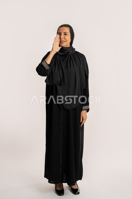 سيدة سعودية تقوم بالنداء بصوت منخفض والكلام بصوت عالي، مفهوم الدردشات والمحادثات، بورتريه مقرب لامرأة عربية خليجية إماراتية محجبة ترتدي عباءة وردية تضع يدها على فمها بإيماءات تدل على المناداة، صورة الجسم بالكامل، خلفية بيضاء