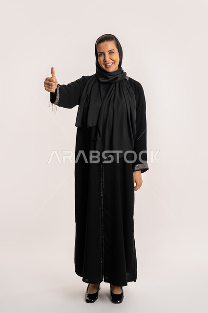 الشعور بالرضى و القبول و الموافقة, النظر الى الكاميرا بتعابير السرور، سيدة سعودية تشعر بالإعجاب، بورتريه لإمراة عربية خليجية اماراتية مبتسمة محجبة ترتدي العباءة السوداء ترفع اصبع الابهام للاعلى بإيماءات علامة الجودة و الامتياز, صورة الجسم بالكامل، خلفية بيضاء