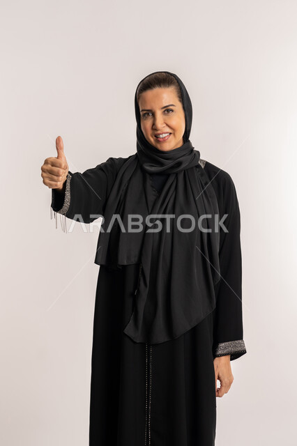 النظر الى الكاميرا بتعابير السرور، سيدة سعودية تشعر بالإعجاب، بورتريه لإمراة عربية خليجية اماراتية مبتسمة محجبة ترتدي العباءة السوداء ترفع اصبع الابهام للاعلى بإيماءات علامة الجودة و الامتياز, الشعور بالرضى و القبول و الموافقة ,خلفية بيضاء