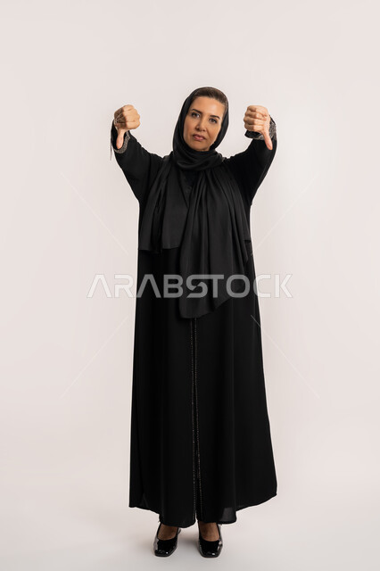 مفهوم الرفض والفشل، سيدة سعودية تشعر بالإنزعاج من شيء ما، بورتريه لإمرأة مسنة عربية خليجية اماراتية ترتدي الحجاب والعباءة تخفض ابهامها بايماءات الرفض وعدم القبول، الاناقة والاهتمام بالمظهر الخارجي، صورة الجسم بالطول الكامل، خلفيه بيضاء