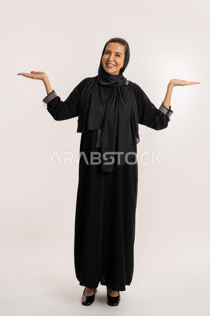 Saudi woman standing with gestures of happiness and joy when seeing someone, gestures indicating reception and welcome, pointing to something, portrait of an elderly Emirati Gulf Arab woman wearing an abaya and hijab raising her arms up with expressions of joy, full body image, white background