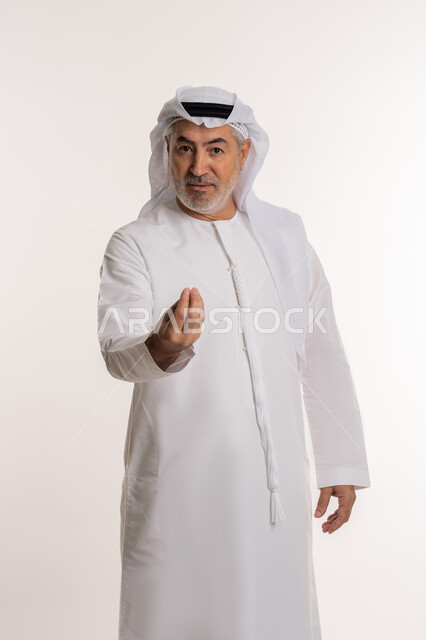Taking some time to do something, concept of patience and deliberation, portrait of an elderly Arab Gulf Emirati man wearing a kandura and a ghutra looking at the camera and making gestures asking to wait, white background