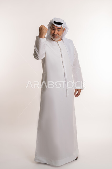 تحقيق الاهداف و إنجاز المهامات, الإعجاب والرضا و الشعور بالسعاده، بورتريه لرجل كبير بالسن عربي اماراتي خليجي يرتدي الكندورة الاماراتية والغترة ويرفع يديه فرحا بالنصر, ايماءات وجه ويدين تدل على الفوز و الكسب, علامة الفرح والقوه, صورة الجسم بالكامل، خلفية بيضاء