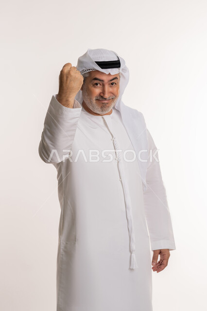 الإعجاب والرضا و الشعور بالسعاده، تحقيق الاهداف و إنجاز المهامات , بورتريه لرجل كبير بالسن عربي اماراتي خليجي يرتدي الكندورة الاماراتية والغترة ويرفع يديه فرحا بالنصر, ايماءات وجه ويدين تدل على الفوز و الكسب, علامة الفرح والقوه, خلفية بيضاء