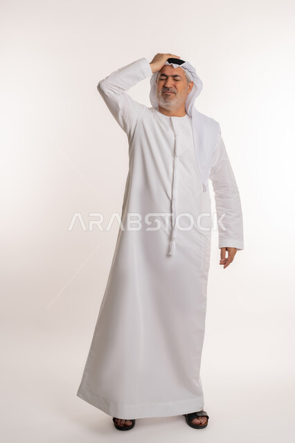 Feeling headache and dizziness, need for treatment and rest, migraine pain, gestures of pain, fatigue and illness, portrait of an elderly Arab Gulf Emirati man wearing a ghutra and kandura, putting his hands on his head feeling dizzy, full body image, white background