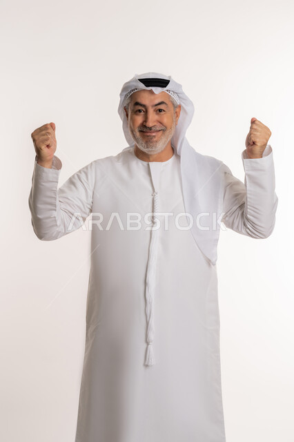 تحقيق الاهداف و إنجاز المهامات, الإعجاب والرضا و الشعور بالسعاده، بورتريه لرجل كبير بالسن عربي اماراتي خليجي يرتدي الكندورة الاماراتية والغترة ويرفع يديه فرحا بالنصر, ايماءات وجه ويدين تدل على الفوز و الكسب, علامة الفرح والقوه, خلفية بيضاء
