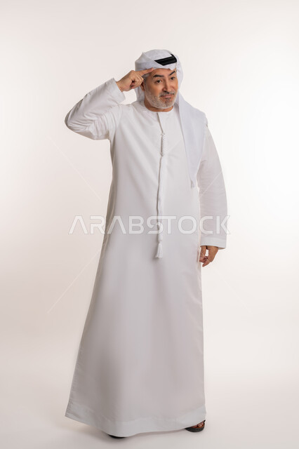 مفهوم السلام والترحيب, تعابير الوجه ولغة الجسد المختلفة, بورتريه لرجل كبير بالسن عربي اماراتي خليجي يرتدي الكندورة الاماراتية والغترة يرفع اليد اليسرى لإلقاء التحية, ايماءات تدل على السعادة, صورة الجسم بالطول الكامل، خلفية بيضاء
