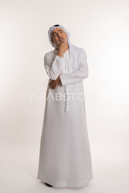 Planning for the future and setting future goals, looking away absentmindedly, portrait of an elderly Arab Gulf Emirati man wearing a kandura and ghutra standing and putting his hand on his chin with gestures of worry and thinking about something, full body image, white background