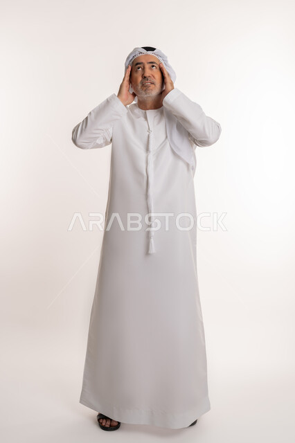الإحساس بوجع الرأس و الدوخة, الحاجة للعلاج والراحة، آلام الشقيقة والصداع النصفي ,إيماءات الألم والتعب والمرض, بورتريه لرجل كبير السن عربي خليجي اماراتي يرتدي الغترة والكندورة يضع يديه على رأسه يشعر بالدوار, صورة الجسم بالكامل، خلفية بيضاء