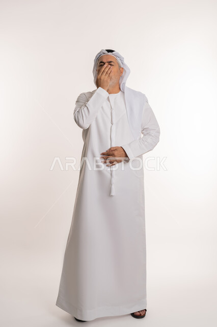 الشعور بالمرض و الزكام والحاجة الى العلاج والدواء, التهاب الأنف التحسسية, بورتريه من الجانب لرجل كبير بالسن عربي اماراتي خليجي يرتدي الغترة والكندورة يضع يديه على وجهه بإيماءات العطاس, صورة الجسم بالكامل، خلفية بيضاء