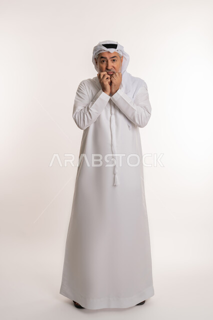 مفهوم الشخصية الانطوائية، شرود الذهن والتفكير والتأمل, حركات يدين ووجه تدل على الخوف والرهبه، بورتريه لرجل كبير بالسن عربي اماراتي خليجي يرتدي الكندورة والغترة الاماراتية يضع يديه تحت ذقنه شعورا بالاحباط و الاستياء، صورة الجسم بالطول الكامل، خلفية بيضاء
