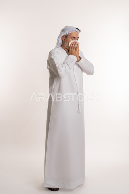 الشعور بالمرض و الزكام والحاجة الى العلاج والدواء, التهاب الأنف التحسسية, استخدام المناديل الورقيه للعطس, بورتريه لرجل كبير بالسن عربي اماراتي خليجي يرتدي الغترة والكندورة يضع يديه على وجهه بإيماءات العطاس, صورة الجسم بالطول الكامل, خلفية بيضاء