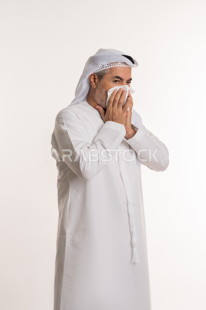 استخدام المناديل الورقيه للعطس, الشعور بالمرض و الزكام والحاجة الى العلاج والدواء, التهاب الأنف التحسسية, بورتريه لرجل كبير بالسن عربي اماراتي خليجي يرتدي الغترة والكندورة يضع يديه على وجهه بإيماءات العطاس, خلفية بيضاء