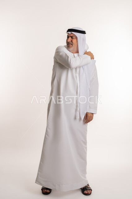 إيماءات الألم في الأكتاف، التهاب العضلات والمفاصل، الإحساس بالتعب والإرهاق, بورتريه لرجل مسن عربي اماراتي خليجي يرتدي الكندورة والغترة يضع يده على كتفه بتعابير الوجع، الحاجه لوضع خطة علاجية, طلب المساعده للذهاب الى المستشفى، صورة الجسم بالكامل، خلفية بيضاء