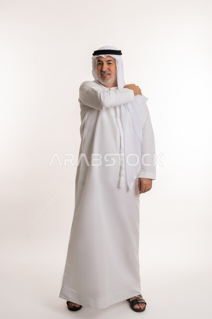 إيماءات الألم في الأكتاف، التهاب العضلات والمفاصل، الإحساس بالتعب والإرهاق, بورتريه لرجل مسن عربي اماراتي خليجي يرتدي الكندورة والغترة يضع يده على كتفه بتعابير الوجع، الحاجه لوضع خطة علاجية, طلب المساعده للذهاب الى المستشفى، صورة الجسم بالكامل، خلفية بيضاء