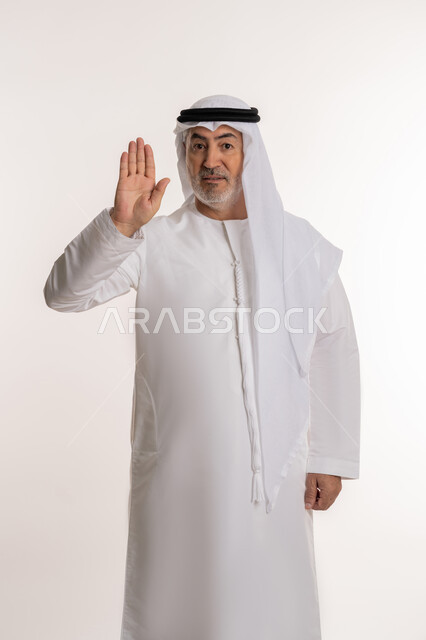 النظر إلى الكاميرا بتعابير الحزم والجدية، طلب المنع والتوقف عن فعل شيء ما، بورتريه لرجل كبير بالسن عربي خليجي إماراتي يرتدي الكندورة التقليدية والغترة يرفع يده بإيماءات الرفض والامتناع، خلفية بيضاء