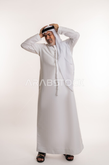 Migraine pain, feeling of headache and dizziness, need for treatment and rest, gestures of pain, fatigue and illness, portrait of an elderly Arab Gulf Emirati man wearing a ghutra and kandura, putting his hands on his head feeling dizzy, full body image, white background