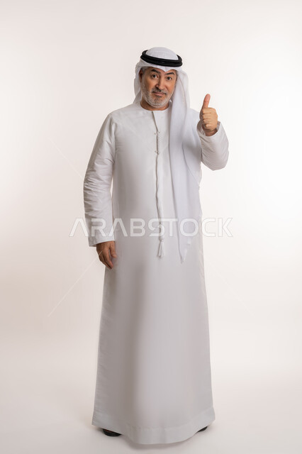 حركات الاعجاب والقبول, النظر إلى الكاميرا بإشارات الموافقه، الشعور بالرضا و السعادة, بورتريه لرجل كبير السن عربي خليجي اماراتي مبتسم يرتدي الكندورة والغترة يرفع يده للأعلى بإيماءات علامة الجودة و السرور, صورة الجسم بالكامل، خلفية بيضاء