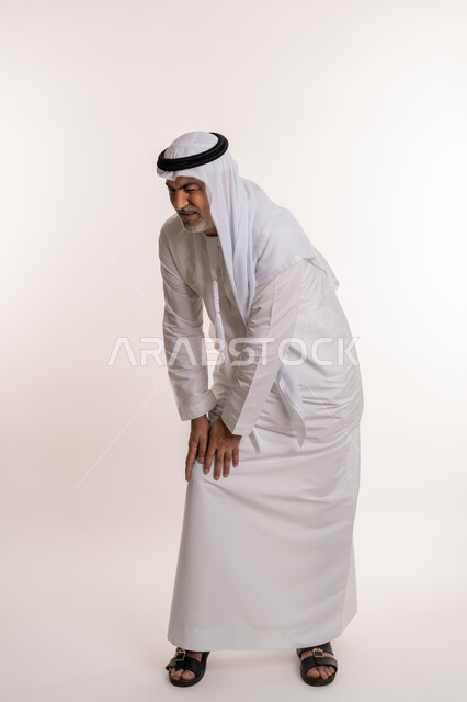 الاحساس بألم في الركبة, بورتريه من الخلف لرجل مسن عربي خليجي اماراتي يرتدي الكندورة و الغترة يشعر بأوجاع في قدمه, أمراض المفاصل و العضلات, التعب والإرهاق و الحاجه للعلاج, طلب المساعده للذهاب الى الطبيب, صورة الجسم بالطول الكامل، خلفية بيضاء