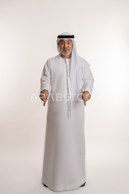 اشارات تحدد هذا المكان (هنا)، تحديد موقع معين, حركات وتعابير وجود شيء ما، بورتريه لرجل عربي خليجي اماراتي كبير بالسن يرتدي الغترة والكندورة يشير بأصابعه نحو الأسفل, النظر إلى الكاميرا بإيماءات السرور، صورة الجسم بالكامل، خلفية بيضاء