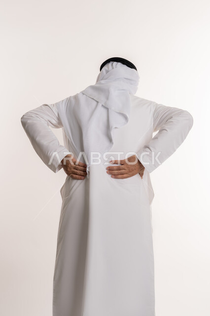 الاحساس بألم في العمود الفقري, بورتريه من الخلف لرجل مسن عربي خليجي اماراتي يرتدي الكندورة و الغترة يشعر بأوجاع في أسفل الظهر, أمراض المفاصل و العضلات, التعب والإرهاق و الحاجه للعلاج, طلب المساعده للذهاب الى الطبيب, خلفية بيضاء