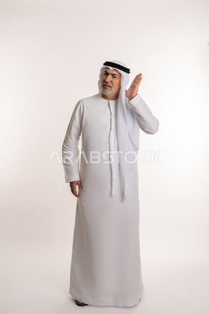 ضعف السمع عند الكبار بالعمر والمسنين، التنصت والتجسس خلسة، الاستماع الى الاسرار والمحادثات, بورتريه لرجل كبير بالسن عربي إماراتي خليجي يضع يده خلف أذنه بايماءات تدل على الانصات، التركيز في الكلام، صورة الجسم بالكامل، خلفيه بيضاء
