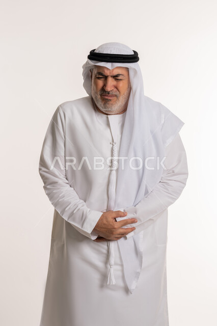 المعاناة من آلام البطن والمعدة، أمراض والتهابات الجهاز الهضمي، الحاجة إلى العلاج، بورتريه لرجل كبير بالسن عربي خليجي إماراتي يرتدي الكندورة والغترة يضع يديه على بطنه بتعابير التعب والإرهاق والإحساس بالمرض،  خلفية بيضاء