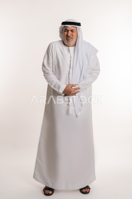 أمراض والتهابات الجهاز الهضمي، الحاجة إلى العلاج، بورتريه لرجل كبير بالسن عربي خليجي إماراتي يرتدي الكندورة والغترة يضع يديه على بطنه بتعابير التعب والإرهاق والإحساس بالمرض، المعاناة من آلام البطن والمعدة، صورة الجسم بالكامل، خلفية بيضاء