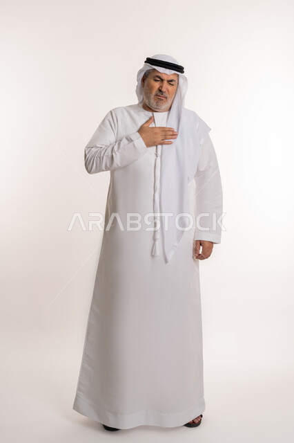 صعوبة التنفس والتعب والألم, الاصابة بنزلة برد والتهاب الحلق، بورتريه لرجل كبير بالسن عربي خليجي إماراتي يرتدي الكندورة والغترة يضع يديه على صدره ويشعر بالاختناق، المرض الشديد والسعال، العدوى بفايروس كورونا، صورة الجسم بالطول الكامل, خلفية بيضاء