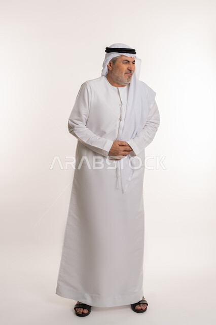 أمراض والتهابات الجهاز الهضمي، الحاجة إلى العلاج، بورتريه لرجل كبير بالسن عربي خليجي إماراتي يرتدي الكندورة والغترة يضع يديه على بطنه بتعابير التعب والإرهاق والإحساس بالمرض، المعاناة من آلام البطن والمعدة، صورة الجسم بالكامل، خلفية بيضاء