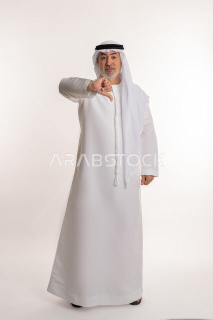 مفهوم الرفض والفشل، الشعور بالإنزعاج من شيء ما، بورتريه لرجل كبير بالسن عربي خليجي اماراتي يرتدي الكندورة والغتره يخفض ابهامه بايماءات الرفض وعدم القبول، الاناقة والاهتمام بالمظهر الخارجي، صورة الجسم بالطول الكامل، خلفيه بيضاء