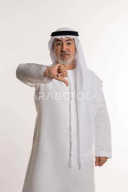 الشعور بالإنزعاج من شيء ما، مفهوم الرفض والفشل، بورتريه لرجل كبير بالسن عربي خليجي اماراتي يرتدي الكندورة والغتره يخفض ابهامه بايماءات الرفض وعدم القبول، الاناقة والاهتمام بالمظهر الخارجي، خلفيه بيضاء