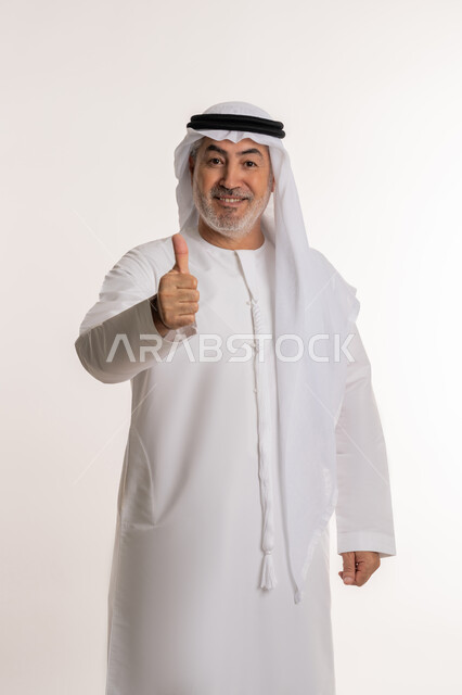 ايماءات القبول والفرح والسرور، الاهتمام بالأناقة والمظهر الخارجي، بورتريه لرجل كبير بالسن عربي خليجي إماراتي يرتدي الكندورة والغترة ينظر الى الكاميرا يرفع إصبع السبابة للاعلى بتعابير الإعجاب والرضا، خلفية بيضاء