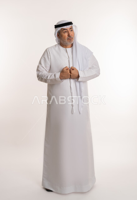 مفهوم الترقب والانتظار، بورتريه لرجل كبير بالسن عربي خليجي إماراتي يرتدي الغترة والكندورة التقليدية يقبض يديه بتعابير التوتر أثناء محاولة إيجاد حل لمشكلة ما، محاولة ضيط الاعصاب والسيطرة على الذات، صورة الجسم بالكامل، خلفية بيضاء