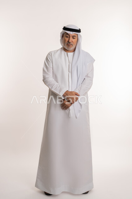شرود الذهن واتخاذ القرارات بتركيز, محاولة إيجاد حل للمشكلات, النظر بتمعن والتخطيط للمستقبل, بورتريه لرجل كبير السن عربي خليجي إماراتي يرتدي الغترة والكندورة يقف بأيدي متماسكة بتعابير العبوس واليأس, الجسم بالطول الكامل, خلفية بيضاء
