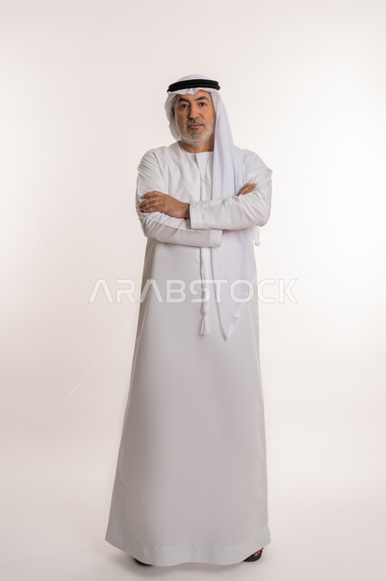 The concept of concern for appearance, masculinity and elegance, looking at the camera with serious and firm gestures, close-up portrait of an elderly Arab Gulf Emirati man wearing a kandura and ghutra, standing with crossed hands with expressions of self-confidence, full-length body image, white background