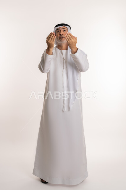 شكر الله وحمده، بورتريه لرجل كبير السن عربي اماراتي خليجي يرتدي الغترة والكندورة التقليدية يرفع يديه بايماءات الدعاء والتضرع، مفهوم طلب العون و المغفرة، الرغبة في تحقيق الأمنيات، صورة الجسم بالطول الكامل, خلفية بيضاء