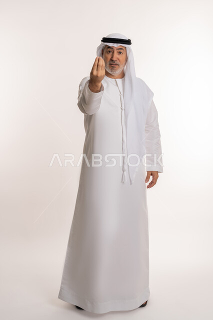 إمهال بعض الوقت للقيام بشئ ما، مفهوم الصبر والتريث، بورتريه لرجل كبير بالسن عربي خليجي اماراتي يرتدي الكندورة والغترة ينظر الى الكاميرا يشير بيده بإيماءات طلب الإنتظار، صورة الجسم بالطول الكامل، خلفية بيضاء