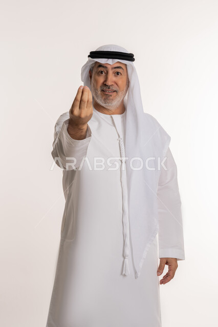 مفهوم الصبر والتريث، إمهال بعض الوقت للقيام بشئ ما، بورتريه لرجل كبير بالسن عربي خليجي اماراتي يرتدي الكندورة والغترة ينظر الى الكاميرا يشير بيده بإيماءات طلب الإنتظار، خلفية بيضاء