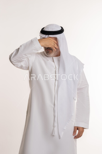 Hand on face and eyes covered, astonishment and surprise at something, remembering something specific suddenly, portrait of an elderly Arab Gulf Emirati man wearing the traditional kandura and ghutra covering his face standing with gestures of astonishment, white background