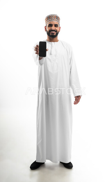 استخدام التكنولوجيا و التقنيات الحديثة المتطورة، بورتريه  لشاب عربي خليجي عماني يرتدي الدشداشه والكمه يقوم بعرض شاشة سوداء فارغة عبر الهاتف المحمول، النظر الى الكاميرا بتعابير السعادة والسرور، خلفية بيضاء