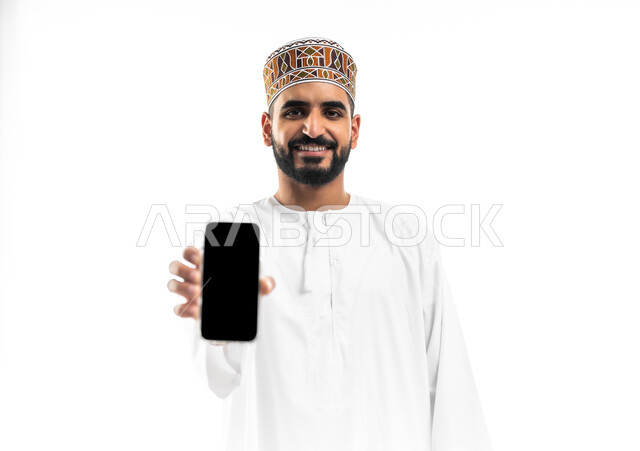 Using advanced modern technology and techniques, portrait of a young Arab Gulf Omani man wearing a dishdasha and a cloak displaying a blank black screen via a mobile phone, looking at the camera with expressions of happiness and pleasure, white background
