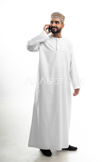 تبادل الأخبار مع الأهل والأصدقاء، إجراء مكالمة هاتفية باستخدام الموبايل، بورتريه لرجل عربي خليجي عماني يرتدي الدشداشة والكمه يقوم بالتواصل مع الأقارب، تعابير الفرح والسرور، خلفية بيضاء