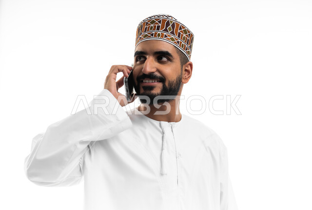 تبادل الأخبار مع الأهل والأصدقاء، إجراء مكالمة هاتفية باستخدام الموبايل، بورتريه لرجل عربي خليجي عماني يرتدي الدشداشة والكمه يقوم بالتواصل مع الأقارب، تعابير الفرح والسرور، خلفية بيضاء