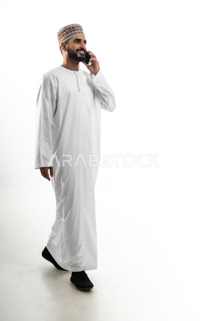 تبادل الأخبار مع الأهل والأصدقاء، إجراء مكالمة هاتفية باستخدام الموبايل، بورتريه لرجل عربي خليجي عماني يرتدي الدشداشة والكمه يقوم بالتواصل مع الأقارب، تعابير الفرح والسرور، خلفية بيضاء