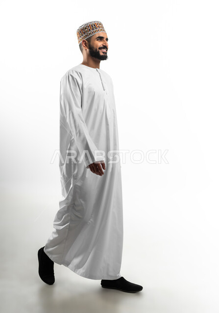 Feeling happy and cheerful, thinking of positive things while walking, walking makes a person happy and pleased, expressions of lightness and psychological comfort, portrait of an Omani young man wearing a dishdasha and a sleeve showing signs of joy, white background