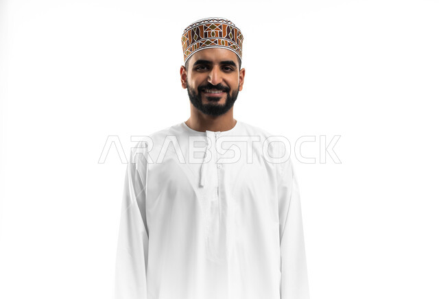 مفهوم النظافة الشخصية، الاهتمام بالمظهر الخارجي، الأناقة في ارتداء الملابس التقليدية، بورتريه لشاب عربي عماني يرتدي الدشداشة والكمه ينظر إلى الكاميرا بتعابير الابتسامة، خلفية بيضاء