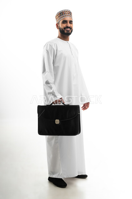 الاهتمام بالأناقة والمظهر الخارجي، الذهاب إلى دوام العمل بنشاط, الشعور بالسرور لتحقيق أهداف الشركة, بورتريه من الجانب لرجل أعمال عربي خليجي عماني مبتسم يرتدي الكمه والثوب التقليدي يحمل حقيبة يد، صورة الجسم بالكامل, خلفية بيضاء