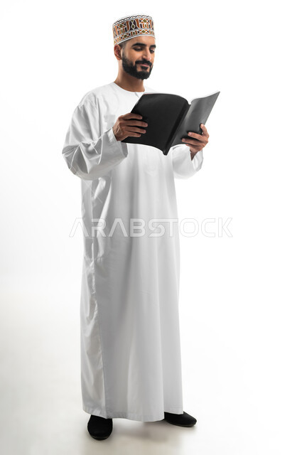 قضاء أوقات الفراغ بالتعلم والتثقف، الاهتمام بتصفح الكتب المفيدة، حب المطالعة والقراءة، بورتريه  لشاب عربي خليجي عماني يرتدي الدشداشه والكمه ينظر الى الكتاب بتمعن وتركيز، ثقافة التعليم الذاتي، خلفية بيضاء